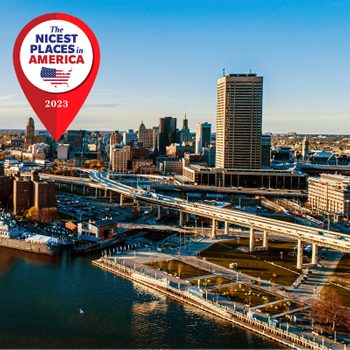 City skyline with roads and riverfront, highlighted by a "Nicest Places in America 2023" pin. Buildings and waterfront park under clear blue sky.