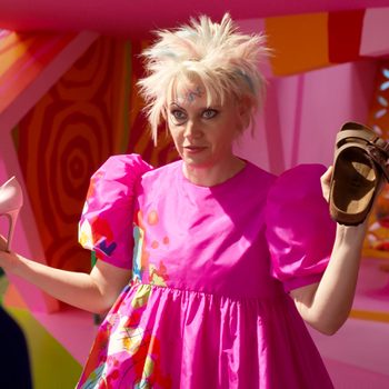 Person holds a pink high heel and a brown sandal, wearing a bright pink dress with puffy sleeves in a colorful, geometric room.