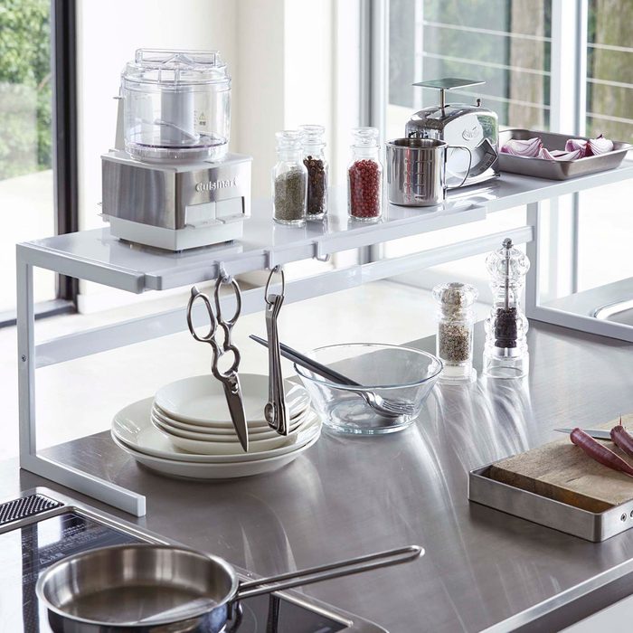 Counter shelf holds kitchen appliances and spices; scissors and measuring spoons hang below. Nearby, dishes, glass bowls, and utensils rest on a metallic surface.