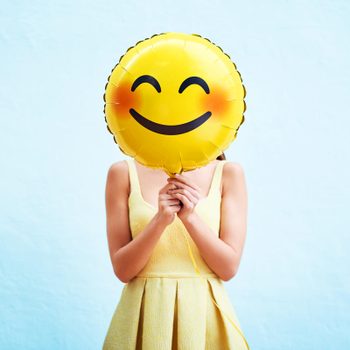 Balloon held covering face, with yellow smiley design, person wearing matching yellow dress, against a light blue background.