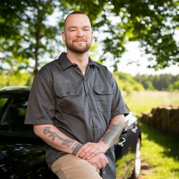 A man stands smiling, arms crossed, in front of a car. He's outdoors, surrounded by greenery, trees, and a stone wall.