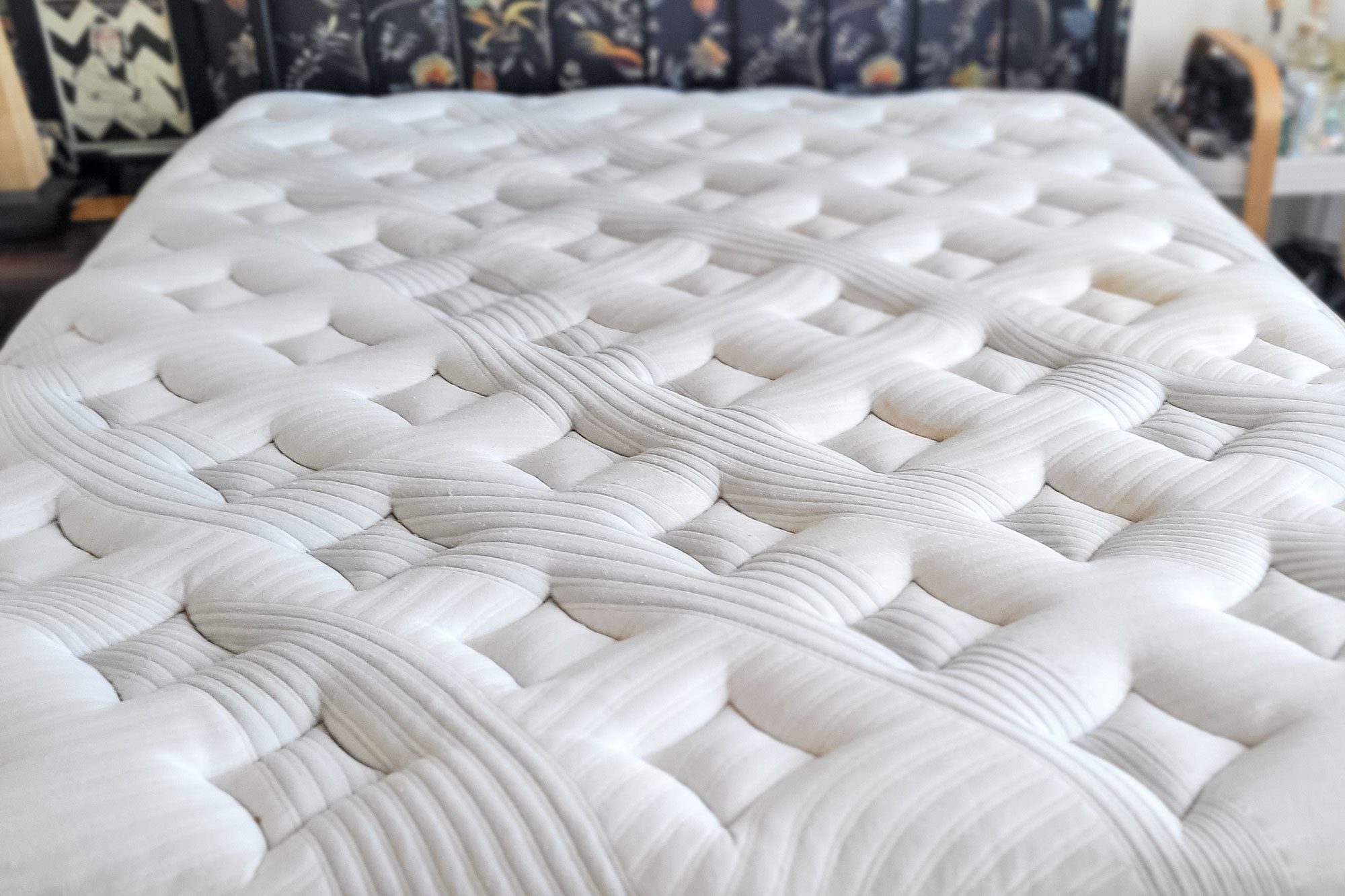A white mattress with a quilted, textured surface lies in a bedroom with patterned wallpaper and wooden furniture.