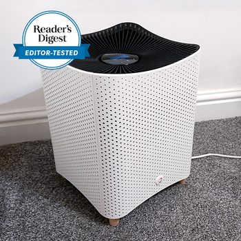 White air purifier operating on gray carpeted floor, featuring a perforated design and digital display. "Readerâ€™s Digest EDITOR-TESTED" badge visible.