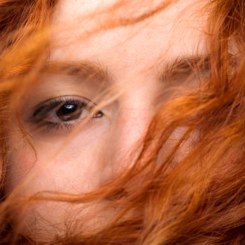 Red hair partially covers a person's eyes and face, with a close-up focus in a soft, natural light ambiance.