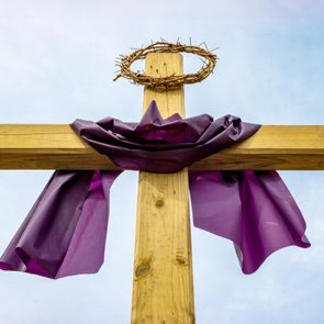 A wooden cross draped with purple cloth stands under a blue sky, topped with a crown of thorns.