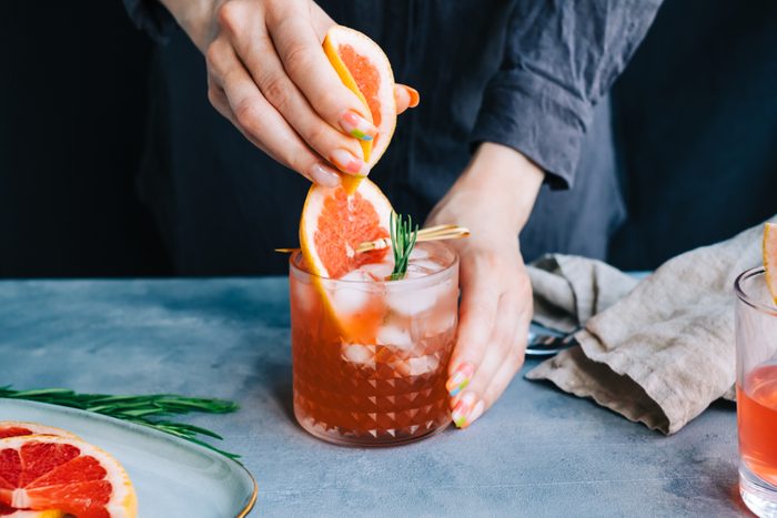 Female bartender hand squeezes juice from fresh grapefruit in cocktail lemonade with ice and rosemary.