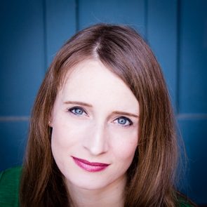 Woman gazes directly, wearing subtle makeup, against a blue background.