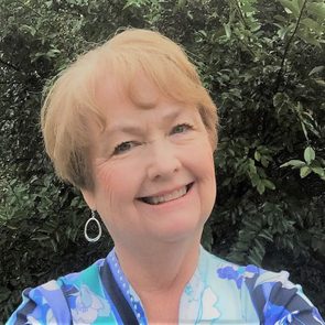 A smiling woman stands outdoors, wearing a floral-patterned garment, with lush green leaves in the background.