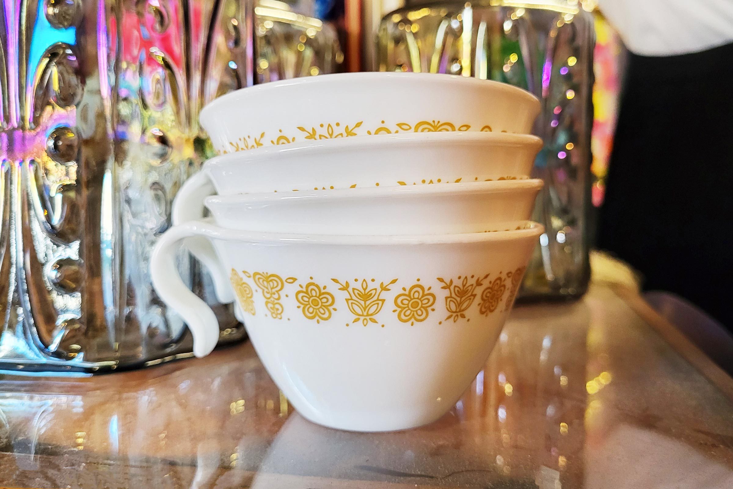 Stacked white cups feature gold floral patterns, resting on a reflective surface beside a colorful, embossed metallic container.
