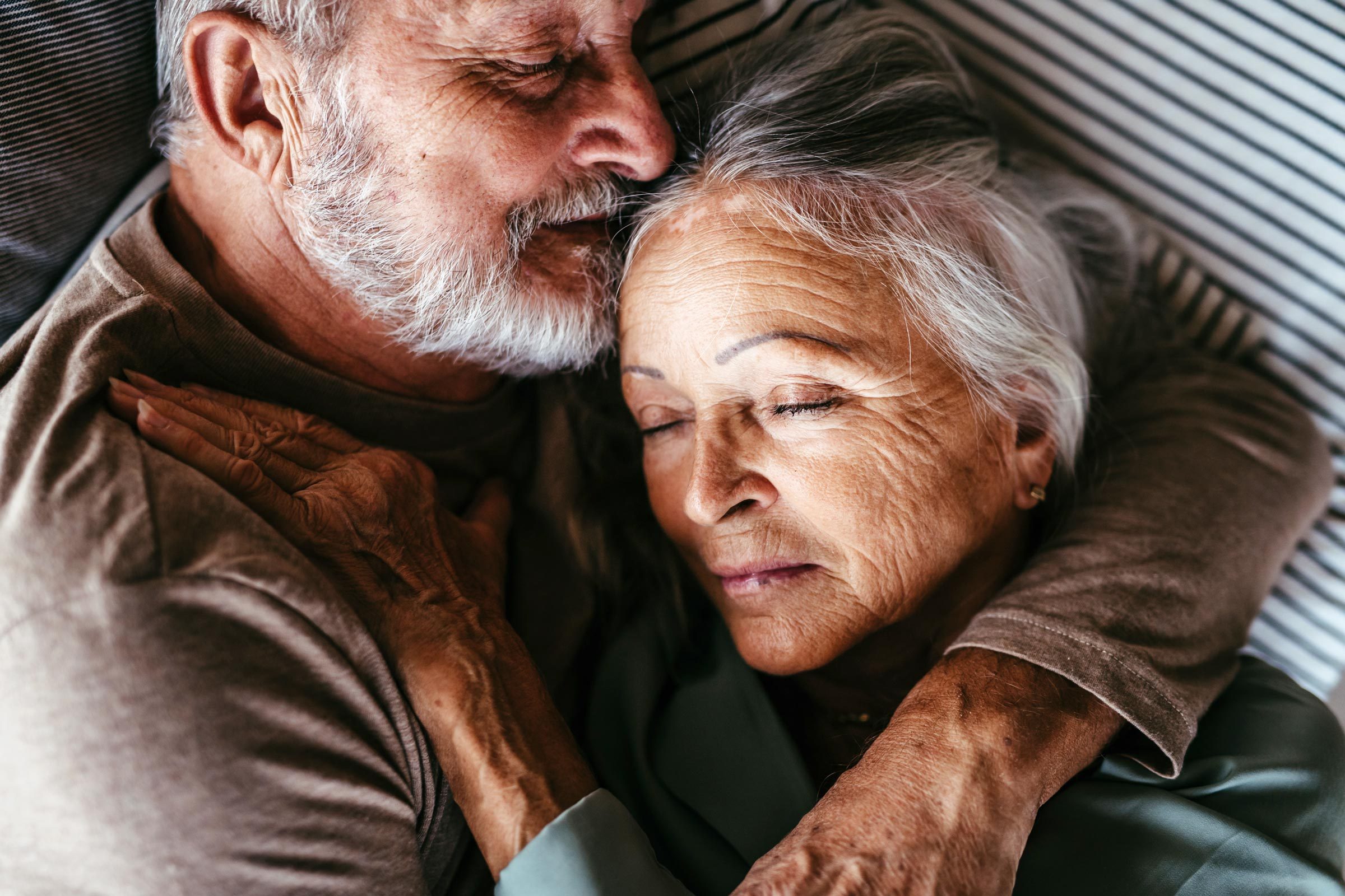Elderly couple cuddling warmly, eyes closed, resting on striped bedding.