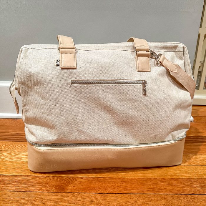A beige canvas bag sits on a wooden floor with a zipper pocket and leather bottom. 
