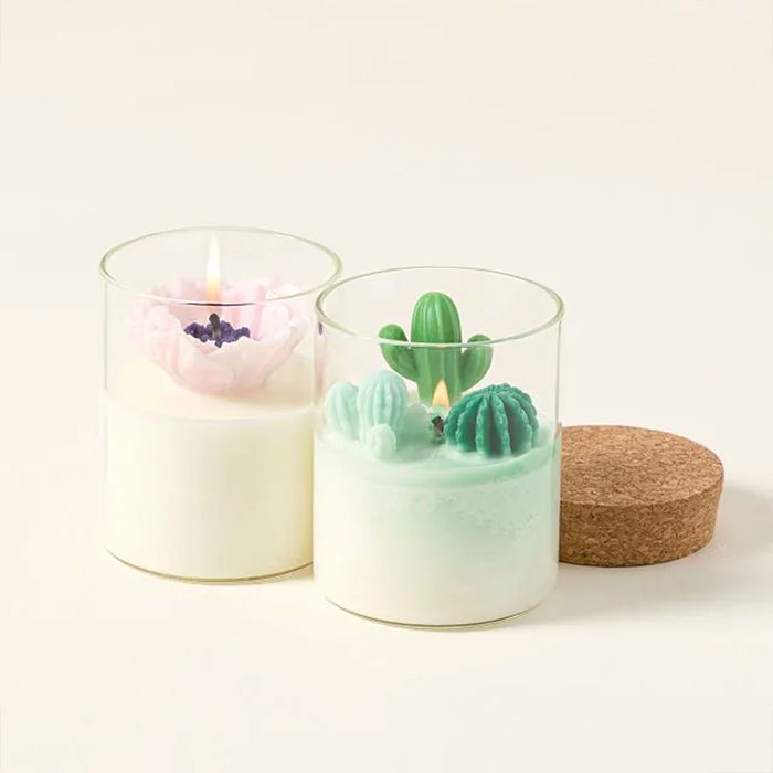 Candles burn softly; one adorned with pink flowers, the other with green cacti designs. They rest on a plain surface with a cork lid nearby.