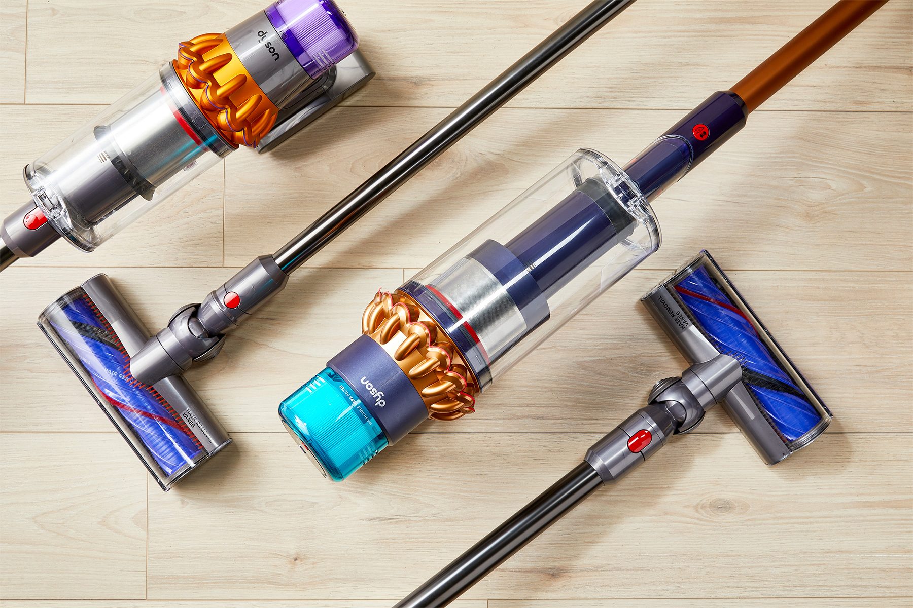 Vacuum cleaners lay on a light wooden floor, showcasing cylindrical canisters, colorful brush heads, and sleek handles in an arranged display.