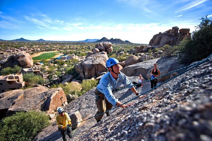 Climbers ascend a rocky slope, wearing helmets and using ropes, with a desert landscape and distant mountains visible in the background under a clear sky.