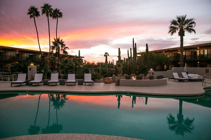 Chairs line a pool beside a crackling fire pit with tall cacti, surrounded by palm trees, under a vibrant sunset sky.