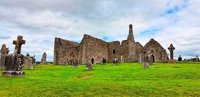 Ruined stone church stands amid gravestones in a green grassy field under a cloudy sky.