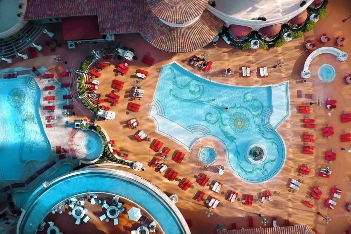 Pools shimmer under sunlight, surrounded by red lounge chairs, within a resort area featuring curved architecture and a terracotta-tiled roof.