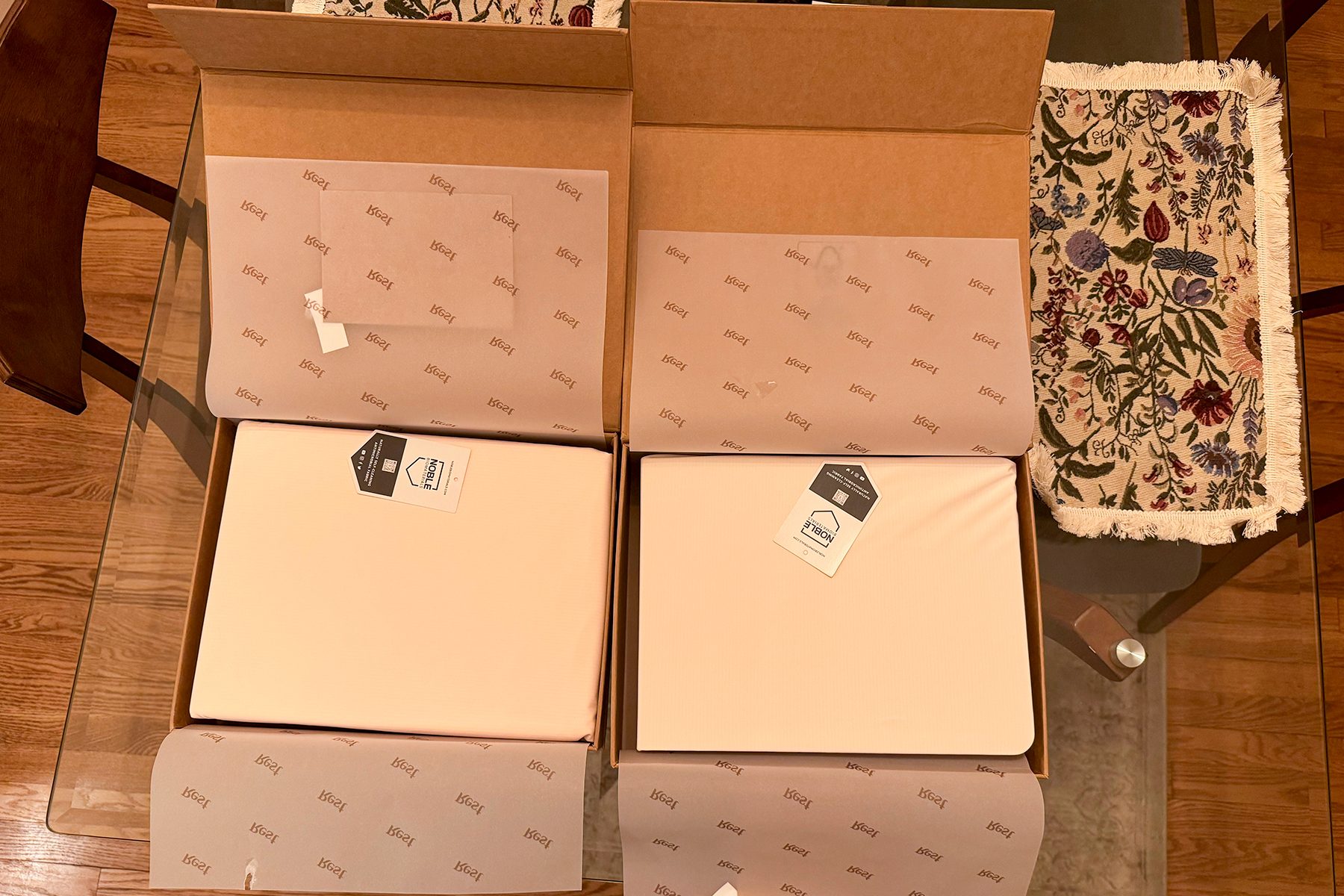 Two cardboard boxes are open on a glass table, containing neatly wrapped items with branded tissue paper and nearby floral-patterned cloth.