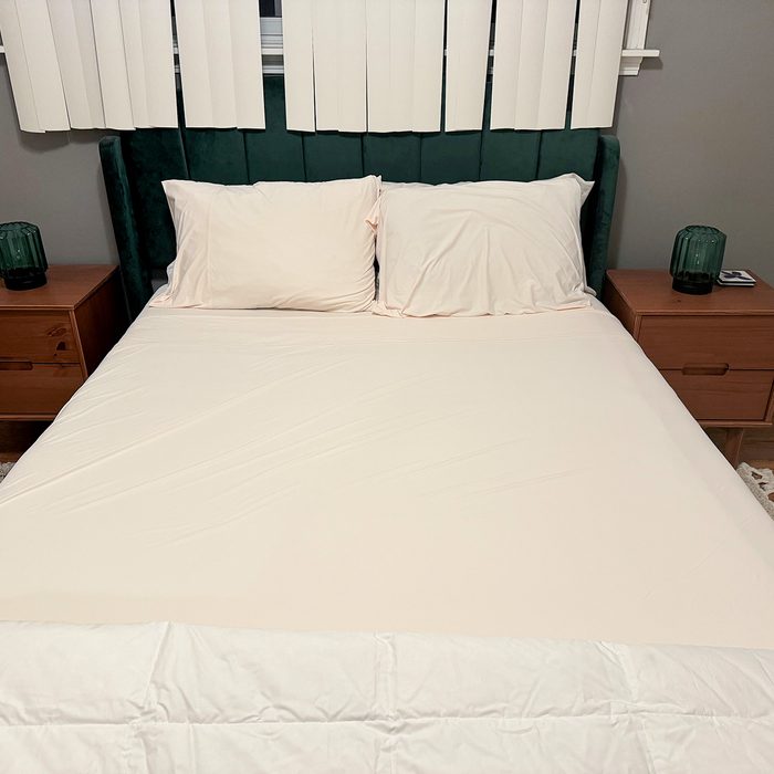 Bed with cream sheets, neatly made, flanked by wooden nightstands with green lamps in a room with vertical window blinds.