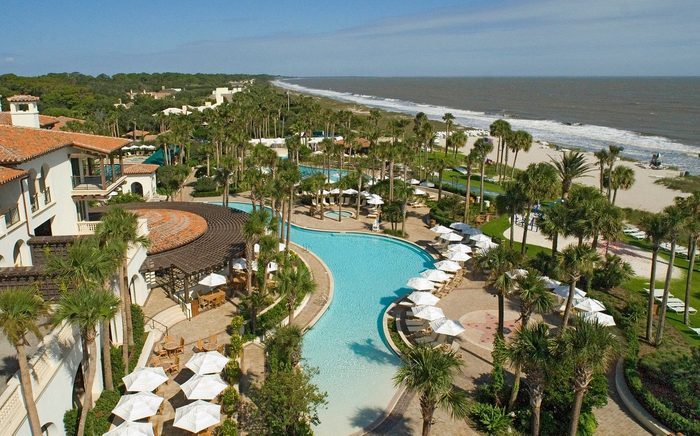 Pool curves through lush resort, surrounded by palm trees and white umbrellas, adjacent to a sandy beach and ocean under a clear blue sky.