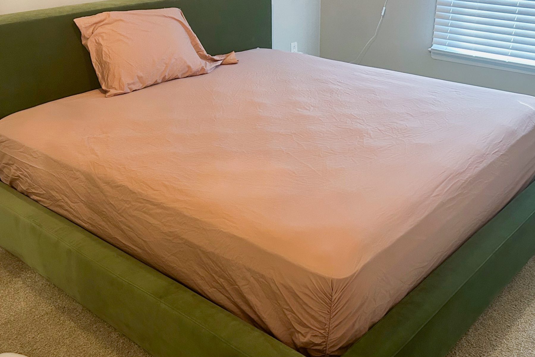 Bed with peach sheets is neatly made on a green frame, in a carpeted room with window blinds partially closed.