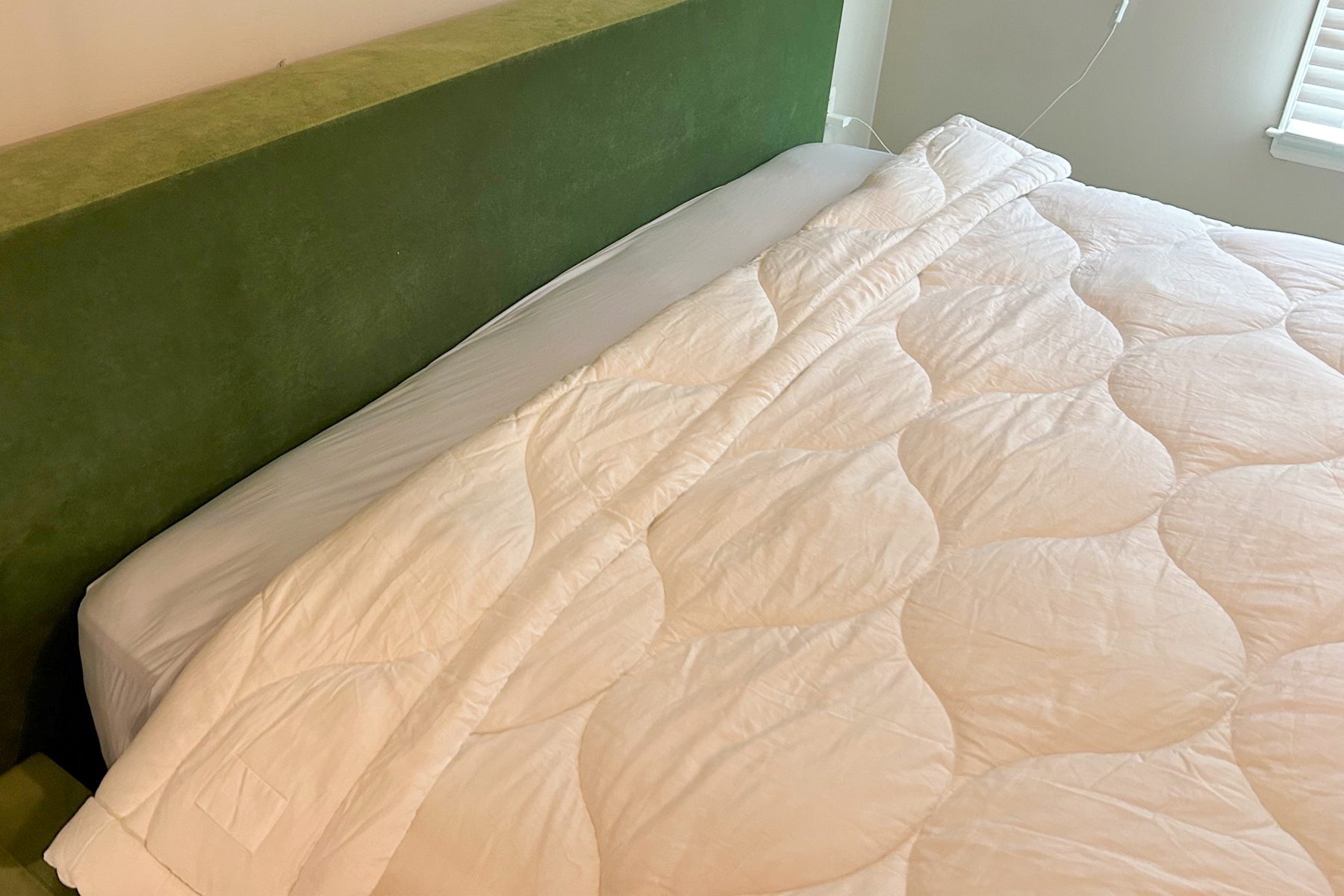 Bed with white, textured comforter neatly spread; green headboard against a light-colored wall; window with blinds partially visible.