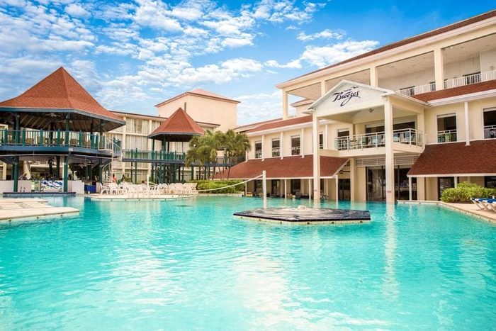 A pool reflects sunshine, surrounded by a resort with multiple buildings and palm trees; a sign reads 