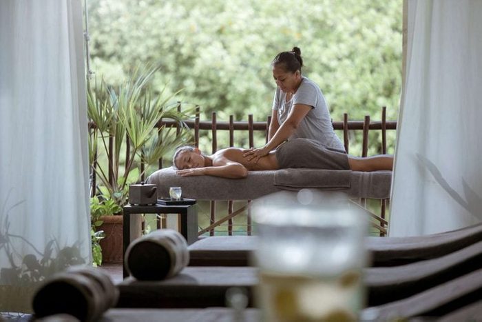 A person receives a massage on a table outdoors, surrounded by plants and draped curtains, with a table holding a candle and a drink nearby.