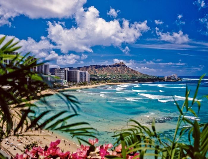 Ocean waves gently reach the shore with lush greenery and vibrant city buildings, under a bright blue sky and scattered clouds.