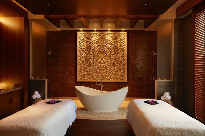 Bathtub centered in luxurious spa room with intricate wall art. Flanked by massage tables, creating a serene, opulent atmosphere. Warm lighting enhances ambiance.