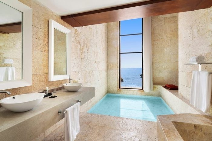 Sink and counter reflect light, positioned in a bright bathroom with large window overlooking ocean, next to a sunlit, stone-walled tub.