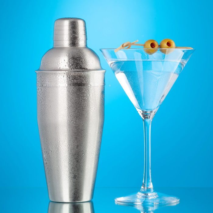 A silver martini shaker stands beside a cocktail in a glass, garnished with olives, against a bright blue background.
