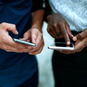 Two hands hold smartphones, interacting actively, in an outdoor setting.