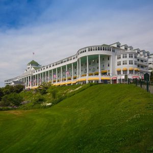 Building extends along a green hillside, displaying American flags under a cloudy sky, with yellow awnings lining the veranda.