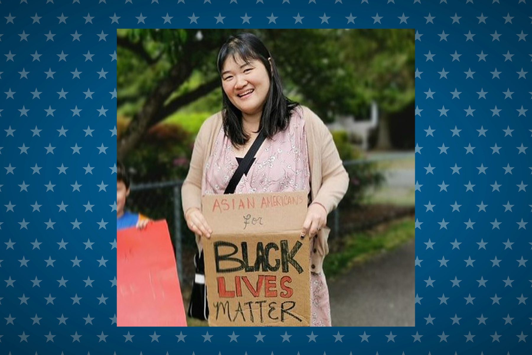 Person holding a sign reading "Asian Americans for Black Lives Matter," standing outdoors with trees in the background, smiling.