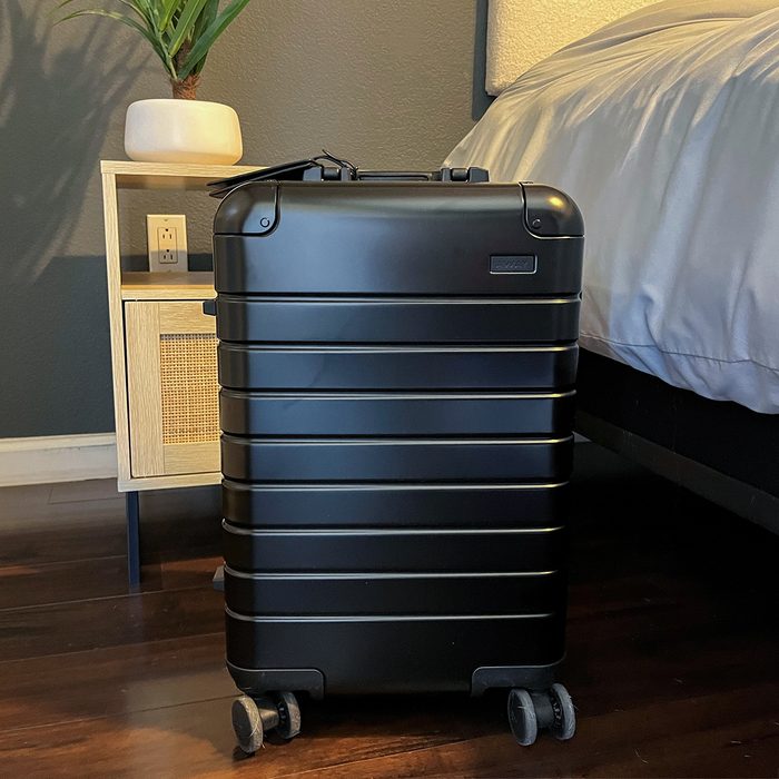 Black suitcase stands beside a nightstand within a bedroom featuring wooden floors and gray walls, showing a potted plant and white bed linens nearby.