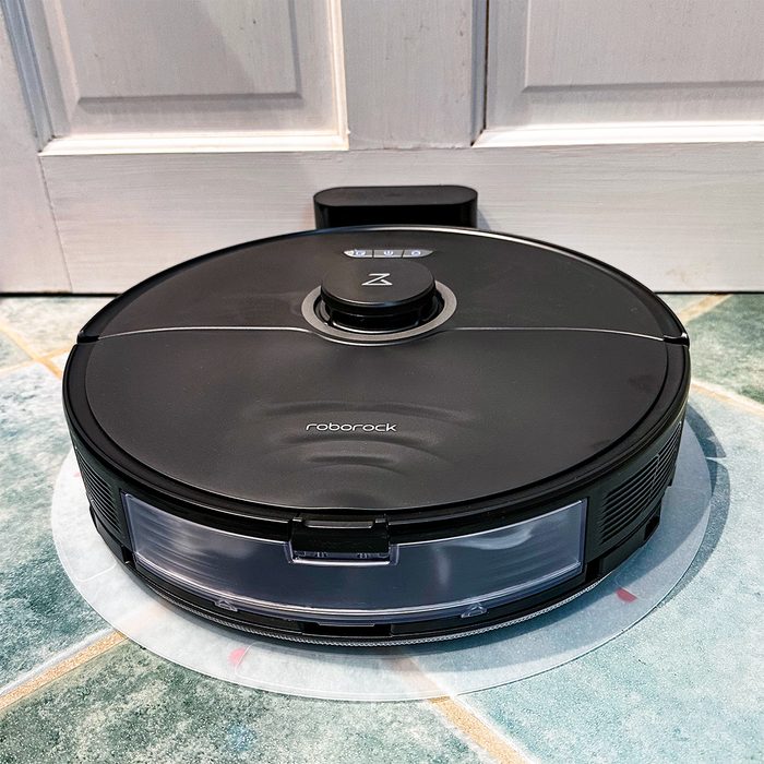 A robotic vacuum sits idly on a tiled floor against a white door, showing the brand name 