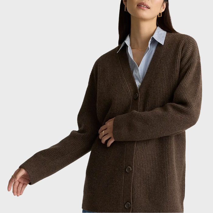 A brown cardigan is worn over a blue striped shirt. The person poses casually in front of a plain background.