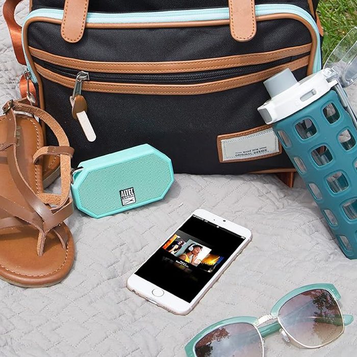 Speaker plays music near sandals, phone, and sunglasses on a quilt; a backpack and water bottle are nearby.