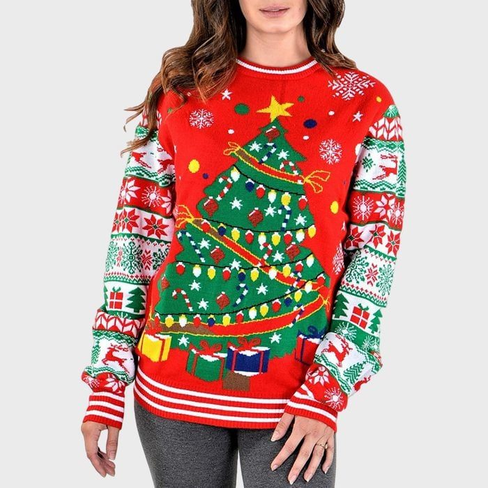 Sweater features colorful Christmas tree design and gifts, with festive patterns on sleeves; worn by a person in a casual setting.