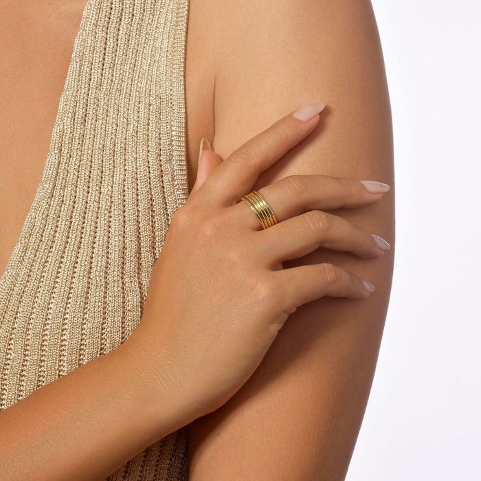 Hand wearing a gold ring rests on an arm, with a textured beige fabric in the background.