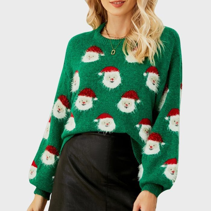 Woman wears a green sweater patterned with Santa faces, paired with a black skirt, standing against a plain gray background.