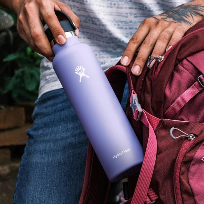 A lavender Hydro Flask is being placed into a red backpack outdoors by a person wearing a patterned shirt and jeans.