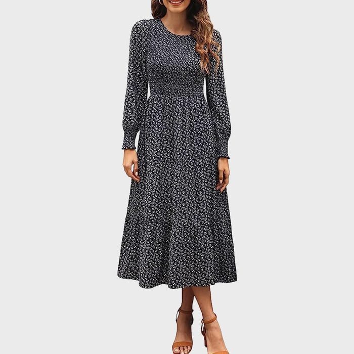Woman wearing a long-sleeved, floral-patterned dress stands smiling against a plain background. Brown sandals are on her feet.