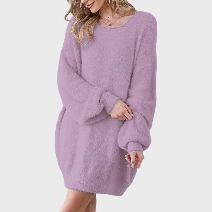 Woman wearing a fluffy, lavender sweater dress, adjusting sleeves. Gray background, relaxed pose, wavy hair partially visible.
