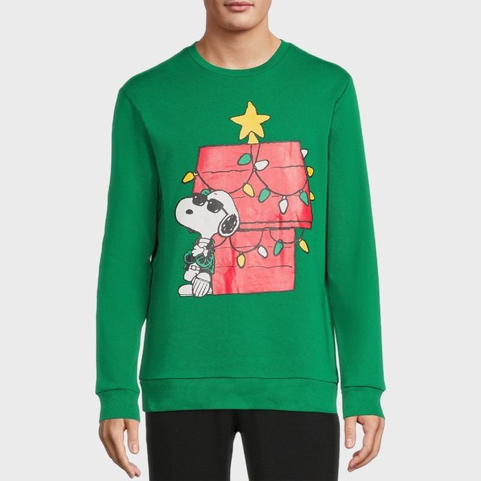 A green sweater displays Snoopy standing beside a decorated, festively lit doghouse, with a yellow star on top, worn by a standing person.