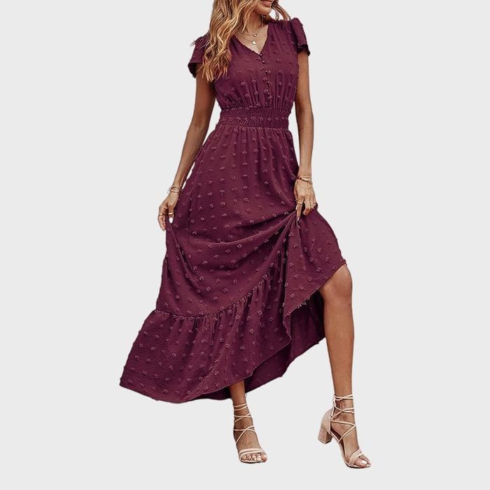 A woman models a maroon dress with a ruffled hem and textured pattern, posing with one hand lifting the skirt, against a plain background.