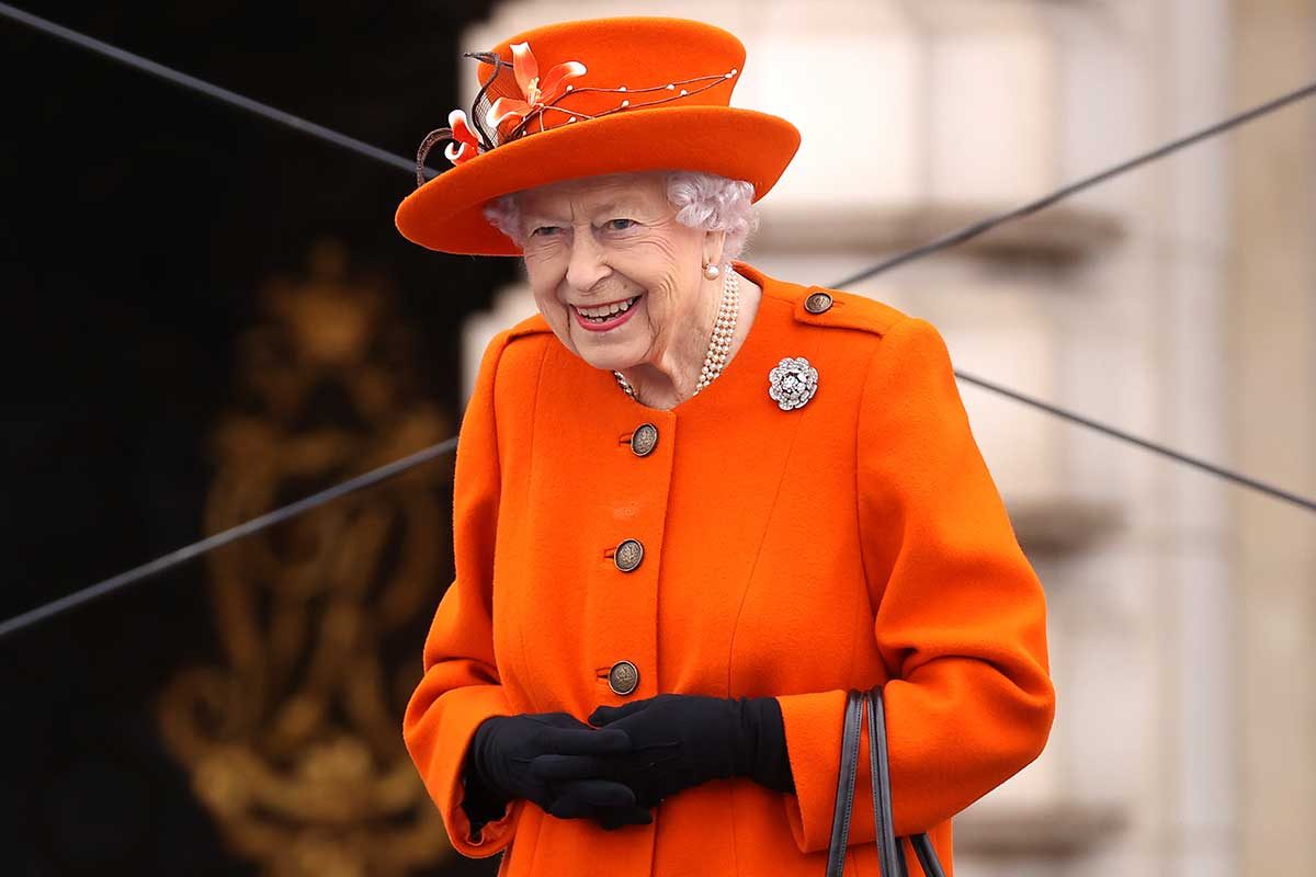 Elderly woman smiling, wearing an orange coat and hat, black gloves, pearl necklace, and brooch; standing outside with blurred architectural background.