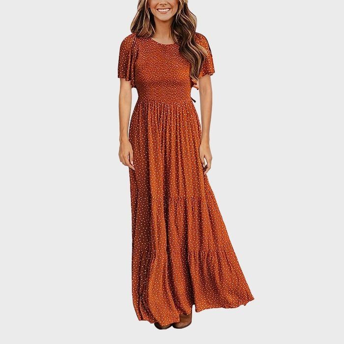 A person stands wearing a long, rust-colored, polka dot dress against a plain white background.