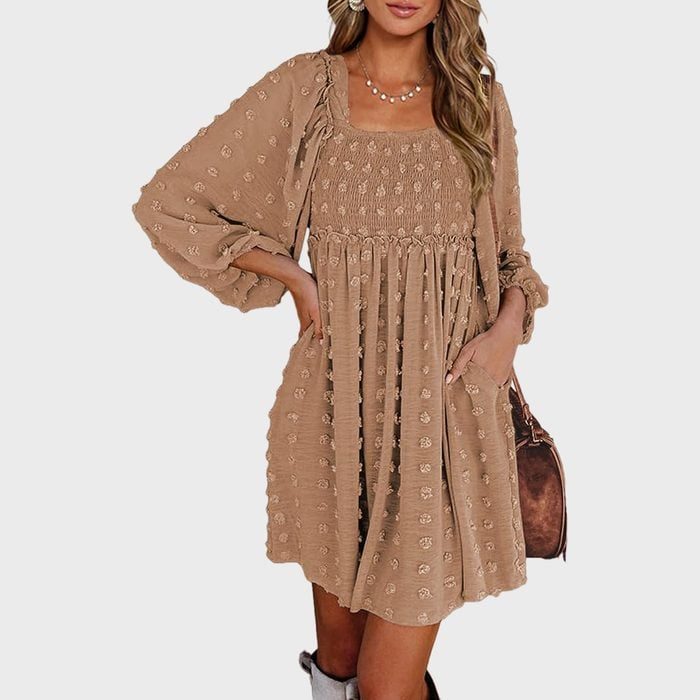 A person models a tan, patterned, long-sleeved dress with a gathered waist, accessorized with jewelry, standing against a plain white background.